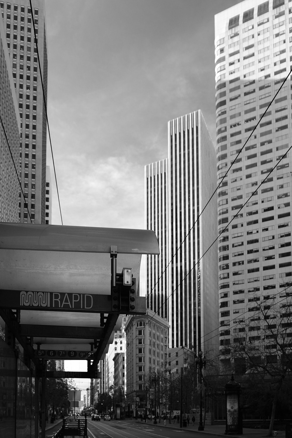 Black and white portrait photo of a MUNI bus stop on Market street surrounded by tall downtown buildings.