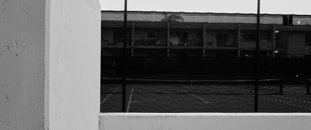 Black and white landscape photo of a tennis court through the black mesh pinned to the high chain-link fence.