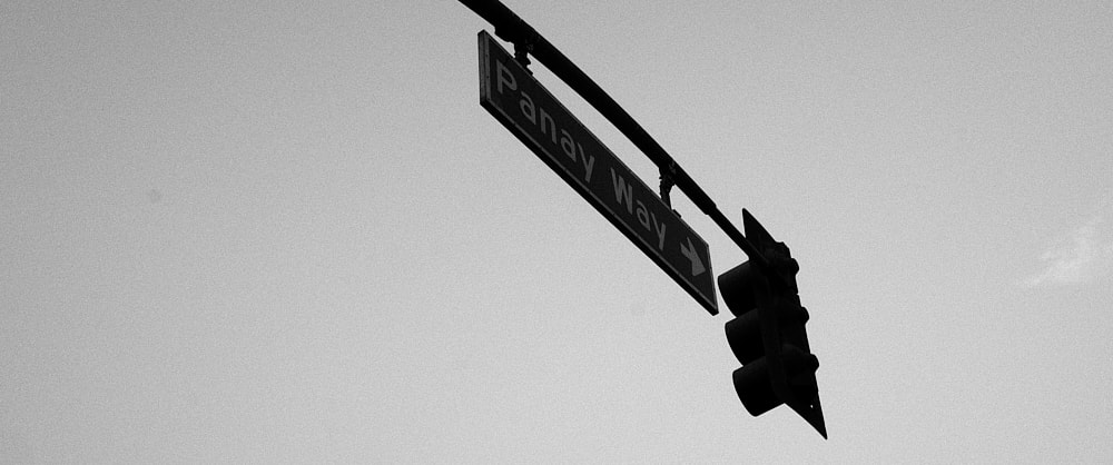 Black and white landscape photo of a traffic signal from behind. A street sign that reads Panaway Way with a white arrow pointing to the right hangs next to the signal.